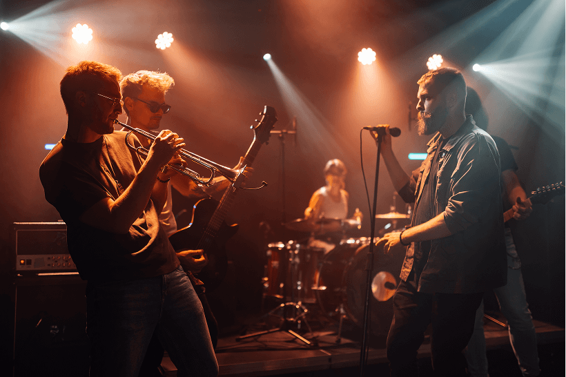 A lively band performs on stage with violin, drums, and guitar under warm lights.