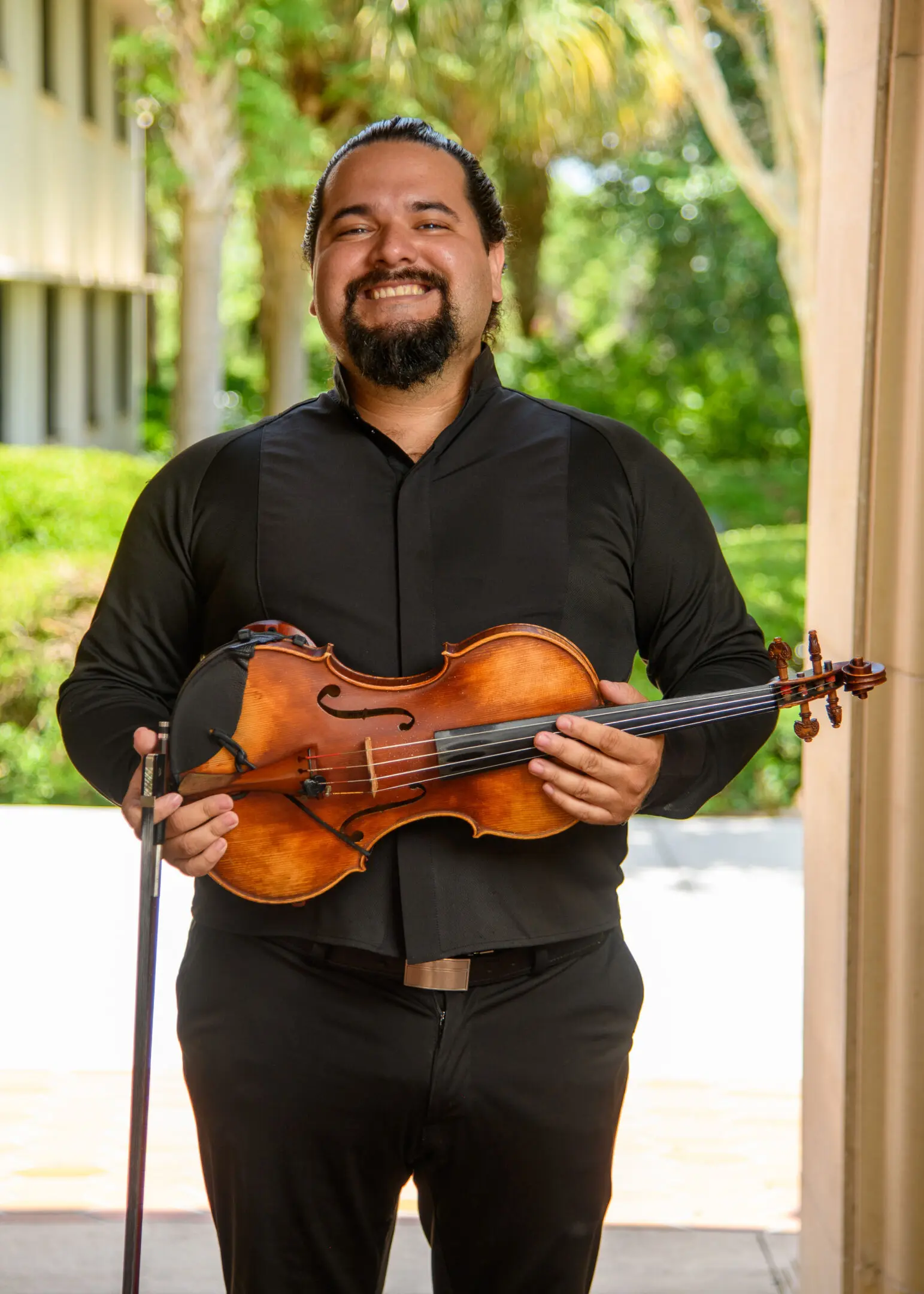 Smiling man holding a violin indoors with greenery outside.