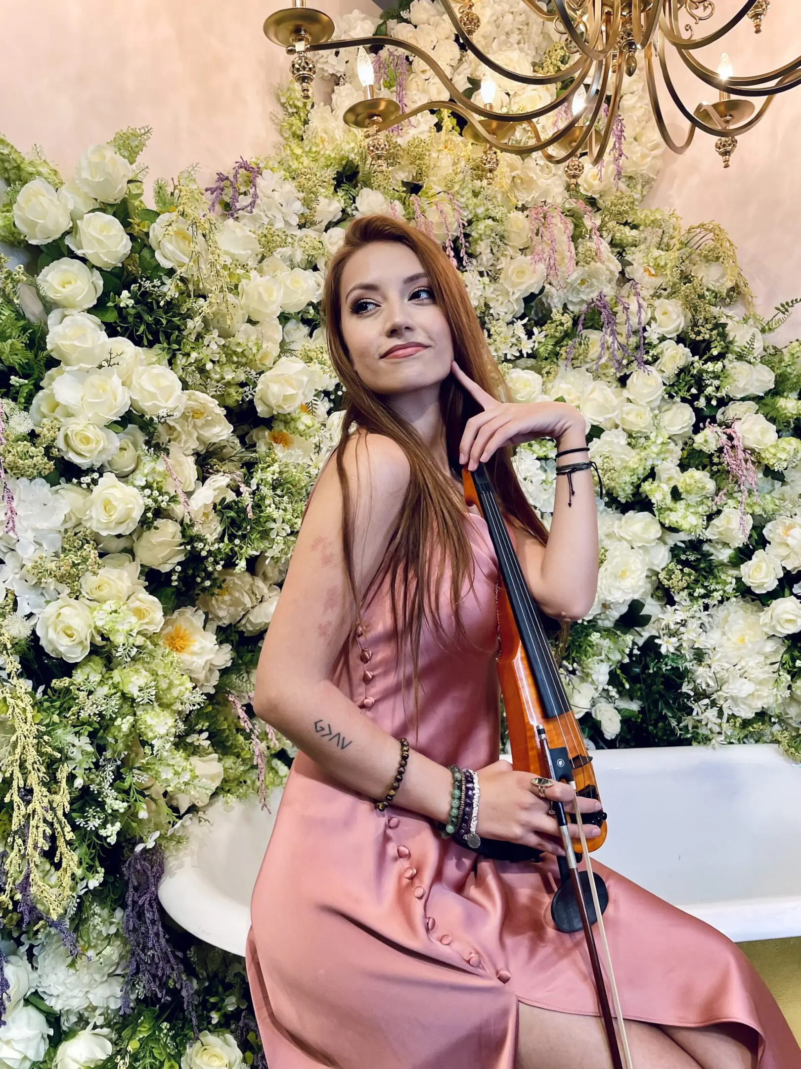 Woman in pink dress posing with violin against floral backdrop.